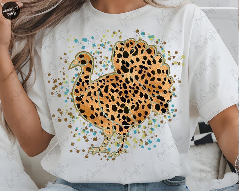 May include: White t-shirt featuring a turkey design with a leopard print pattern. The turkey is outlined in gold and surrounded by colorful paint splatters. The shirt has short sleeves and a crew neck.