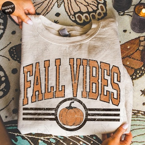 May include: Cream-colored sweatshirt with the text "FALL VIBES" in orange, featuring a pumpkin graphic. The design includes a distressed, vintage aesthetic. The sweatshirt is folded and held by hands.