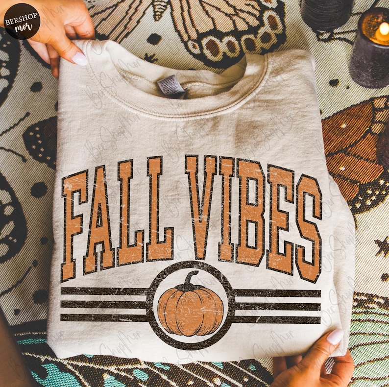 May include: Cream-colored sweatshirt with the text "FALL VIBES" in orange, featuring a pumpkin graphic. The design includes a distressed, vintage aesthetic. The sweatshirt is folded and held by hands.