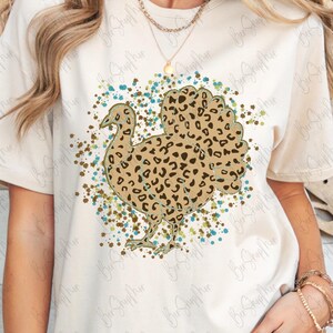 May include: Cream-colored t-shirt featuring a turkey design with a leopard print pattern. The turkey is surrounded by a splatter of teal, brown, and green paint. The shirt has short sleeves and a crew neck.