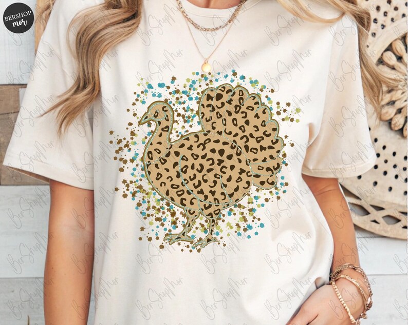 May include: Cream-colored t-shirt featuring a turkey design with a leopard print pattern. The turkey is surrounded by a splatter of teal, brown, and green paint. The shirt has short sleeves and a crew neck.