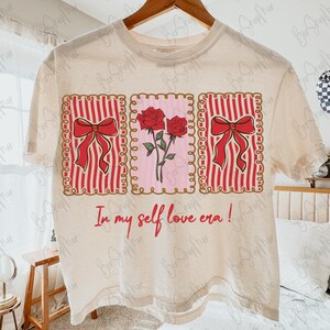 May include: Cream-colored t-shirt featuring three rectangular designs. The left and right rectangles have red bows on red and white striped backgrounds. The center rectangle has red roses on a pink background. The text "In my self love era!" is below.