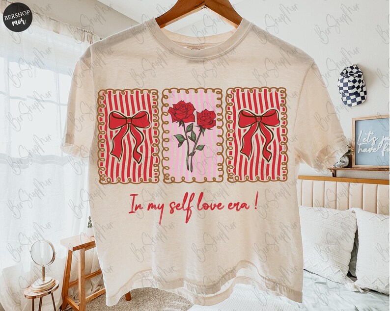 May include: Cream-colored t-shirt featuring three rectangular designs. The left and right rectangles have red bows on red and white striped backgrounds. The center rectangle has red roses on a pink background. The text "In my self love era!" is below.