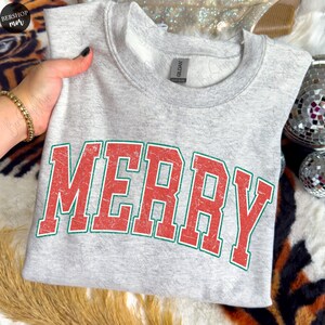 May include: A light gray heathered crewneck sweatshirt featuring the word MERRY printed in large, distressed red block letters with a thin green outline. The garment has a standard ribbed collar and a visible brand tag inside the neckline.