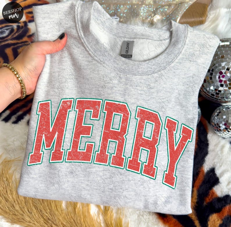 May include: A light gray heathered crewneck sweatshirt featuring the word MERRY printed in large, distressed red block letters with a thin green outline. The garment has a standard ribbed collar and a visible brand tag inside the neckline.