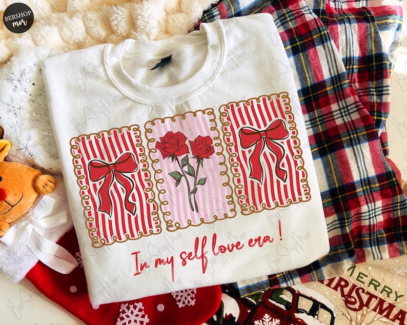 May include: White sweatshirt with a graphic design featuring red bows, roses, and the text "In my self love era!". The design is framed with a gold border. The sweatshirt is part of a holiday theme.