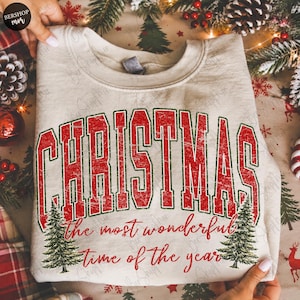 Op de afbeelding: Een beige sweater met ronde hals voorzien van de rode, verweerde tekst CHRISTMAS in een collegiaal lettertype, gevolgd door de rode cursieve tekst the most wonderful time of the year, met twee groene dennenboomillustraties onderaan.
