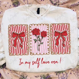May include: White sweatshirt with a graphic design featuring red roses, red bows, and vertical red stripes. The text "In my self love era!" is written in red script below the design. The sweatshirt has a V-neck.