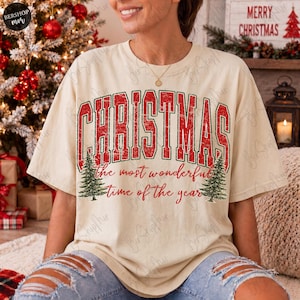 Op de afbeelding: Een beige t-shirt met korte mouwen en een rood-groene opdruk met de tekst CHRISTMAS the most wonderful time of the year. Het ontwerp bevat twee groene dennenbomen aan weerszijden van de tekst, alles met een verweerde textuur.