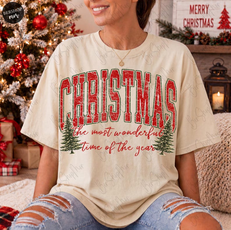 Op de afbeelding: Een beige t-shirt met korte mouwen en een rood-groene opdruk met de tekst CHRISTMAS the most wonderful time of the year. Het ontwerp bevat twee groene dennenbomen aan weerszijden van de tekst, alles met een verweerde textuur.