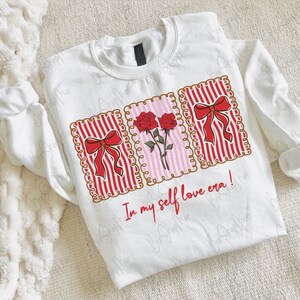 May include: White sweatshirt with a graphic design featuring red bows, roses, and the text "In my self love era!". The design is framed within decorative borders. The long-sleeved top is made of a soft material.