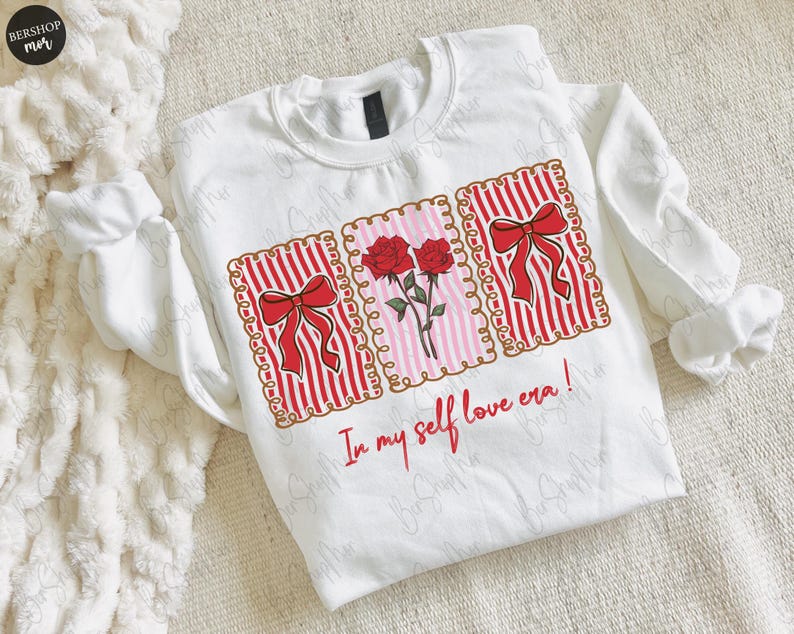 May include: White sweatshirt with a graphic design featuring red bows, roses, and the text "In my self love era!". The design is framed within decorative borders. The long-sleeved top is made of a soft material.