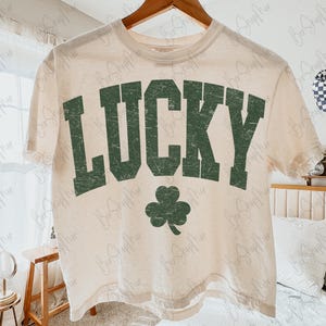 以下が含まれることがあります： 色あせた緑色のブロック文字で「LUCKY」という文字と緑色のクローバーがプリントされた、クリーム色のTシャツ。木製のハンガーにかかっています。