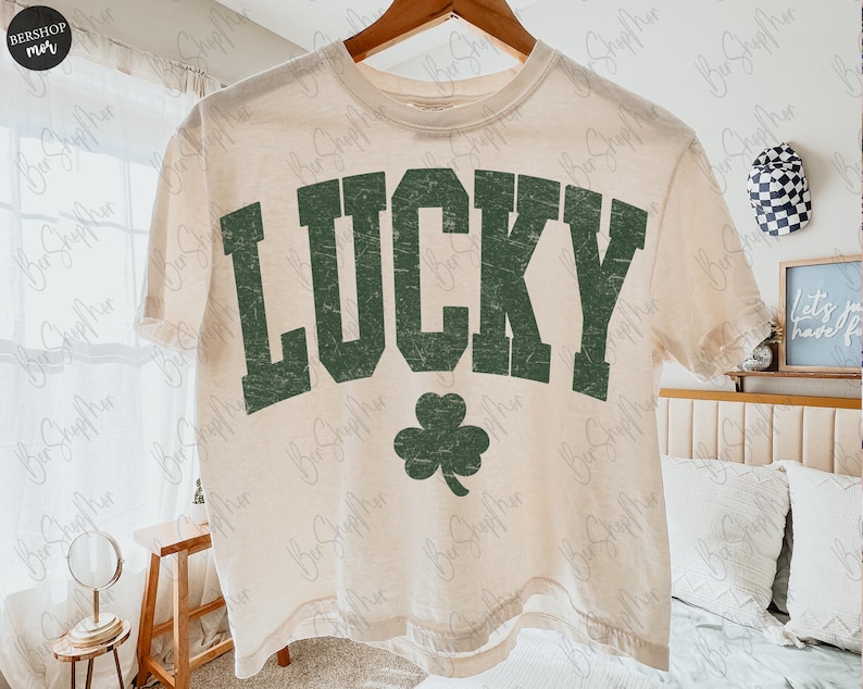以下が含まれることがあります： 色あせた緑色のブロック文字で「LUCKY」という文字と緑色のクローバーがプリントされた、クリーム色のTシャツ。木製のハンガーにかかっています。