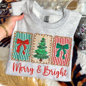 May include: A light gray sweatshirt with a festive design. The design features three rectangular panels with a red bow, a Christmas tree, and a green bow. The words "Merry & Bright" are printed in red script below the panels.