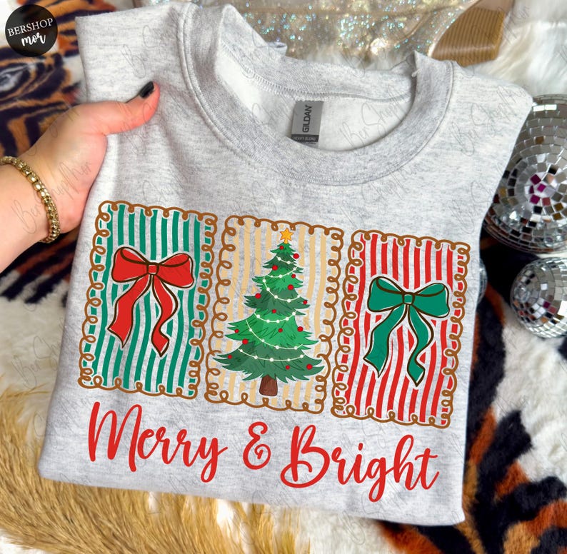 May include: A light gray sweatshirt with a festive design. The design features three rectangular panels with a red bow, a Christmas tree, and a green bow. The words "Merry & Bright" are printed in red script below the panels.