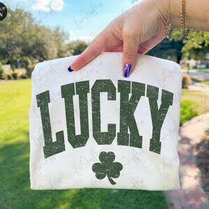 以下が含まれることがあります： 白地のTシャツに、かすれた緑色の文字で「LUCKY」の文字と緑色のクローバーのグラフィックがプリントされています。緑の草と木を背景に、Tシャツが掲げられています。