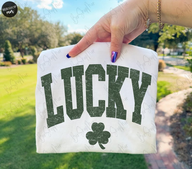 以下が含まれることがあります： 白地のTシャツに、かすれた緑色の文字で「LUCKY」の文字と緑色のクローバーのグラフィックがプリントされています。緑の草と木を背景に、Tシャツが掲げられています。