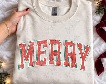 Op de afbeelding: Een crèmekleurig sweatshirt met ronde hals en het woord MERRY in grote, verweerde rode blokletters met een dunne groene omlijning. Het kledingstuk heeft een geribbelde kraag en lange mouwen, uitgevoerd in een zachte, textuurrijke stof.