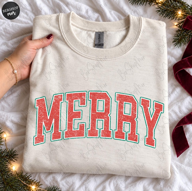 May include: A cream-colored crewneck sweatshirt featuring the word MERRY in large, distressed red block letters with a thin green outline. The garment has a ribbed collar and a standard long-sleeve design, presented on a soft, textured fabric.