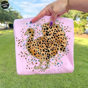 May include: A light pink sweatshirt featuring a turkey design with a leopard print pattern. The turkey is outlined in gold and has black spots. The sweatshirt is decorated with colorful paint splatters.