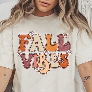 May include: Cream-colored t-shirt with the words "FALL VIBES" in a retro font, decorated with small daisy flowers. The text is in shades of orange, beige, and burgundy. A tattoo of a bicycle is visible on the arm.