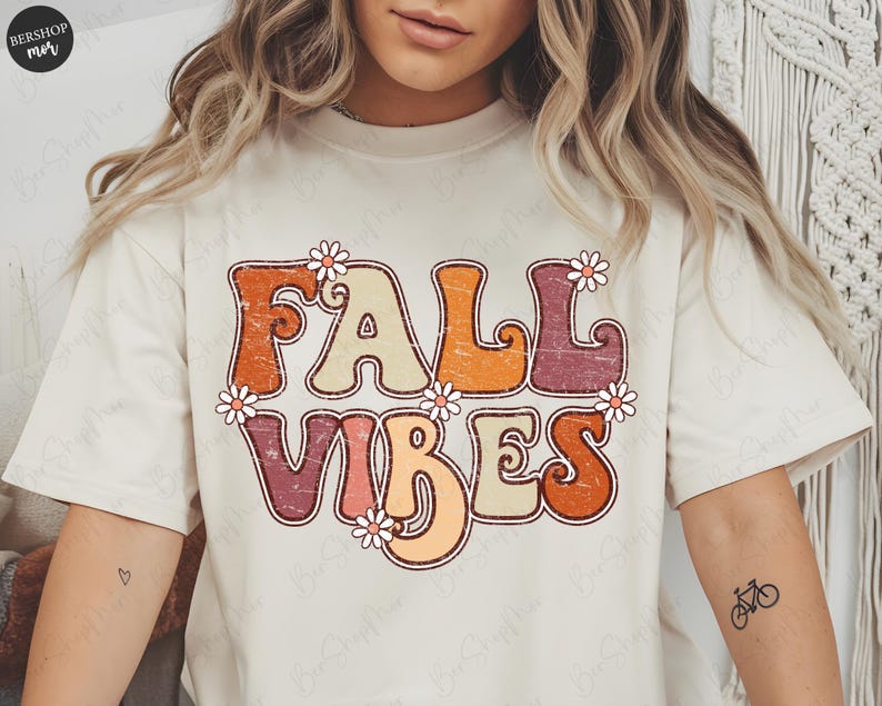 May include: Cream-colored t-shirt with the words "FALL VIBES" in a retro font, decorated with small daisy flowers. The text is in shades of orange, beige, and burgundy. A tattoo of a bicycle is visible on the arm.