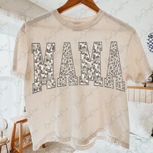 May include: A beige t-shirt with the word "MAMA" printed across the front. The letters have a camouflage and leopard print design. The shirt is hanging on a wooden hanger.