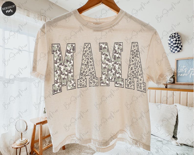 May include: A beige t-shirt with the word "MAMA" printed across the front. The letters have a camouflage and leopard print design. The shirt is hanging on a wooden hanger.