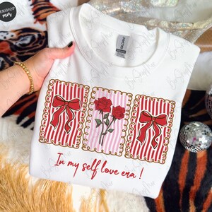 May include: White sweatshirt with a graphic design featuring red bows, roses, and the text "In my self love era!". The design includes red and white stripes and a pink background. The sweatshirt is held by a person.