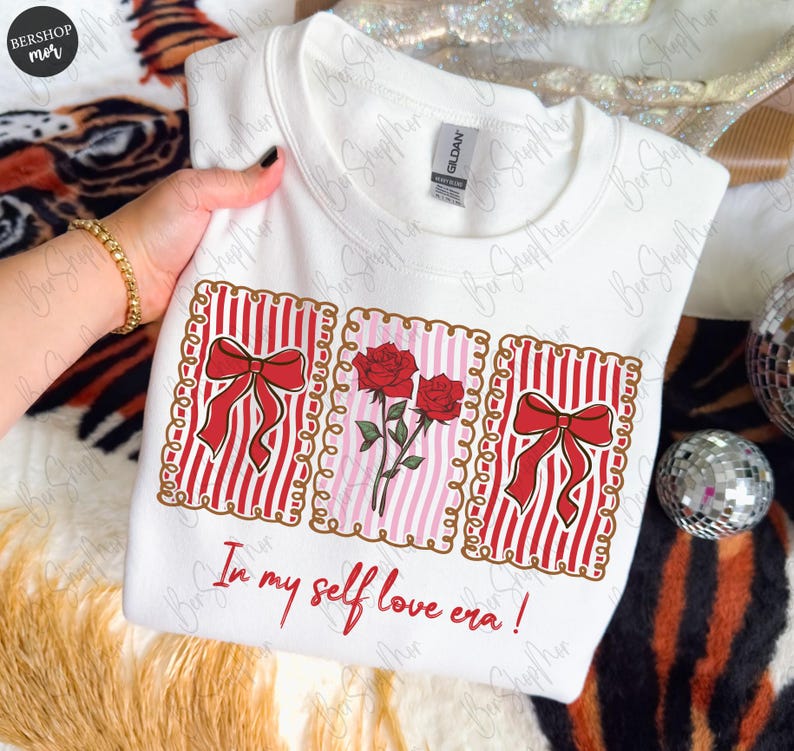 May include: White sweatshirt with a graphic design featuring red bows, roses, and the text "In my self love era!". The design includes red and white stripes and a pink background. The sweatshirt is held by a person.