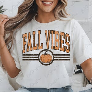 May include: White t-shirt with the text "FALL VIBES" in orange, with a pumpkin graphic. The design includes a distressed, vintage style. The shirt has short sleeves and a crew neck.