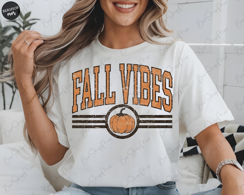 May include: White t-shirt with the text "FALL VIBES" in orange, with a pumpkin graphic. The design includes a distressed, vintage style. The shirt has short sleeves and a crew neck.