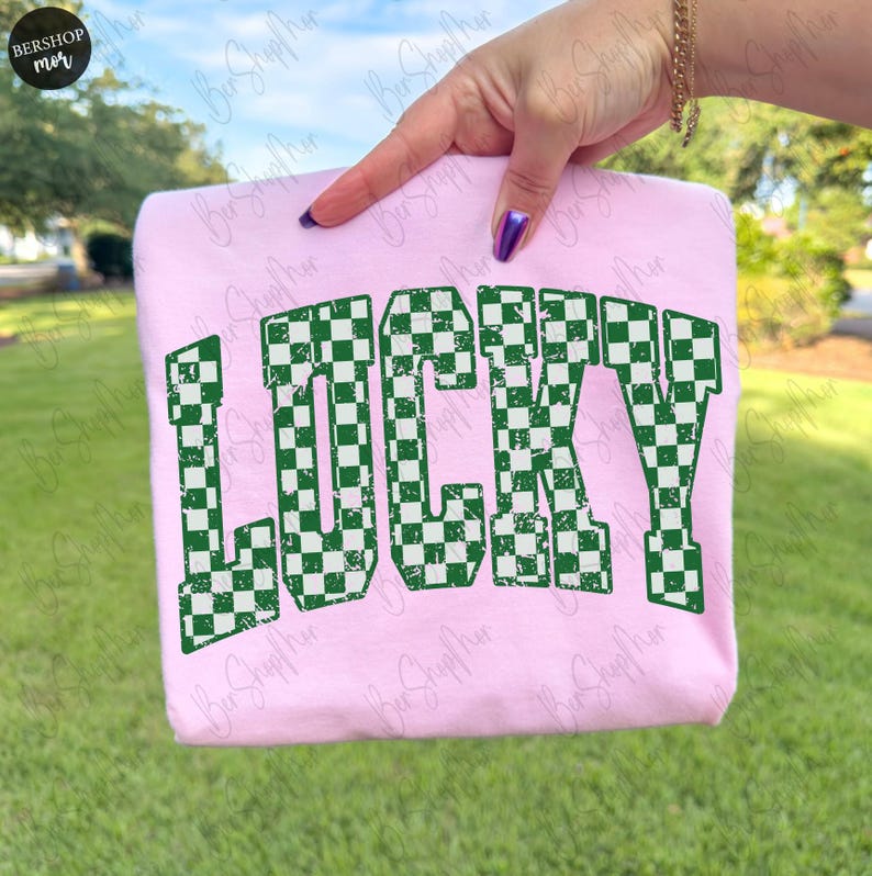 May include: A light pink sweatshirt with the word "LUCKY" in large, distressed, green and white checkered letters. The sweatshirt is held up by a hand with purple nail polish, against a background of green grass and trees.