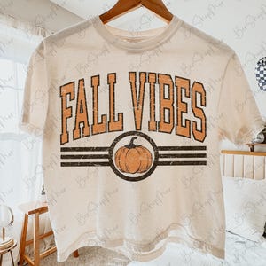 May include: Cream-colored t-shirt with the words "FALL VIBES" in orange block letters. A pumpkin graphic is centered below the text, within a black circle and two black stripes. The shirt is hanging on a wooden hanger.