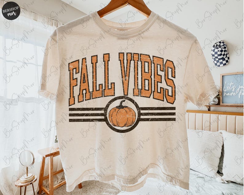 May include: Cream-colored t-shirt with the words "FALL VIBES" in orange block letters. A pumpkin graphic is centered below the text, within a black circle and two black stripes. The shirt is hanging on a wooden hanger.