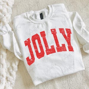 May include: A white sweatshirt with the word "JOLLY" printed in distressed red lettering. The long-sleeved top is made of a soft material and is laid flat on a textured surface. The sleeves are cuffed at the wrists.