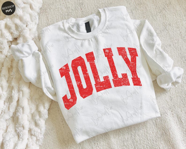 May include: A white sweatshirt with the word "JOLLY" printed in distressed red lettering. The long-sleeved top is made of a soft material and is laid flat on a textured surface. The sleeves are cuffed at the wrists.