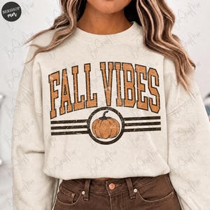 May include: Cream-colored sweatshirt with the text "FALL VIBES" in orange block letters. A pumpkin graphic is centered below the text, within a circle and two horizontal lines. The sweatshirt is a fall-themed apparel item.