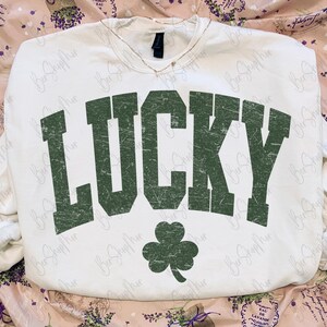 以下が含まれることがあります： 「LUCKY」の文字が緑色でかすれたデザインの白いスウェットシャツと緑色のクローバー。スウェットシャツはVネックで長袖です。背景は花柄です。