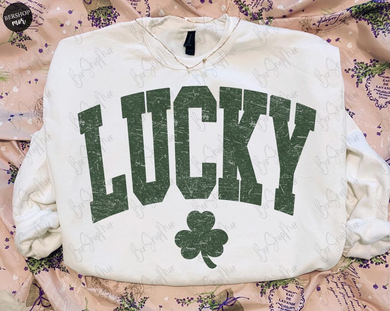以下が含まれることがあります： 「LUCKY」の文字が緑色でかすれたデザインの白いスウェットシャツと緑色のクローバー。スウェットシャツはVネックで長袖です。背景は花柄です。