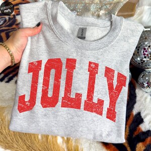 May include: A light gray sweatshirt with the word "JOLLY" printed in distressed red block letters. The crew neck sweatshirt is made of a soft material and is perfect for the holiday season.