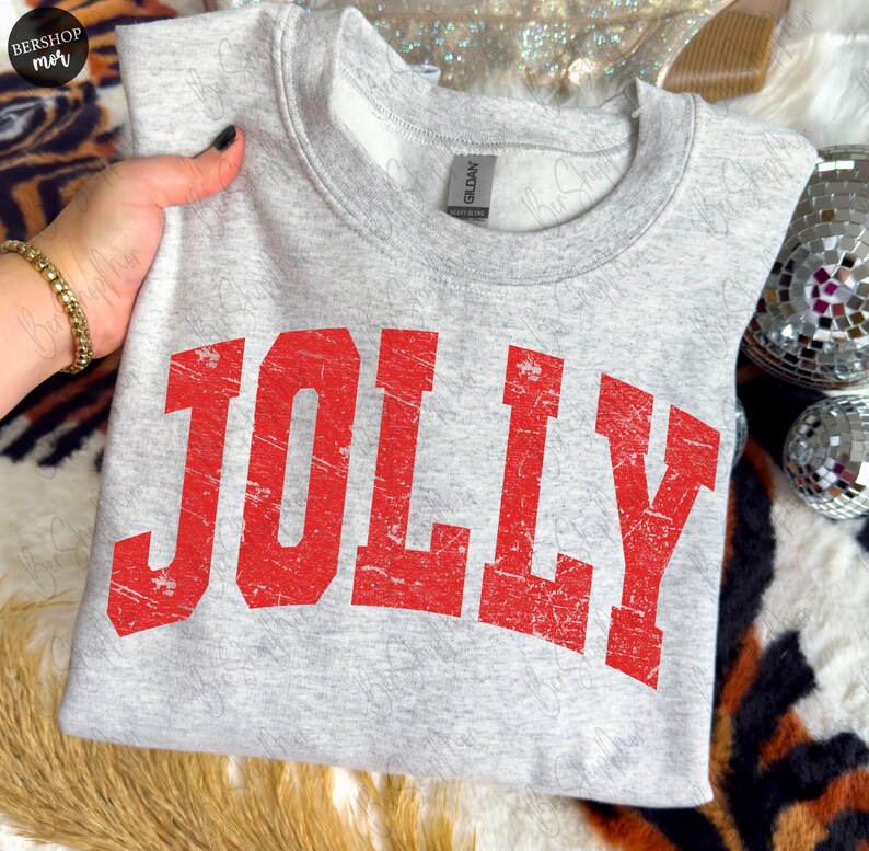 May include: A light gray sweatshirt with the word "JOLLY" printed in distressed red block letters. The crew neck sweatshirt is made of a soft material and is perfect for the holiday season.