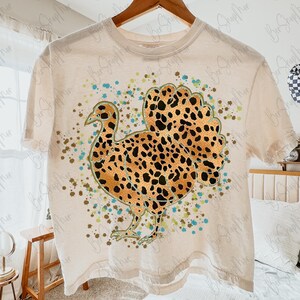 May include: A cream-colored t-shirt featuring a turkey design with a leopard print pattern on the body and tail. The turkey is surrounded by teal and brown speckles. The shirt is hanging on a wooden hanger.