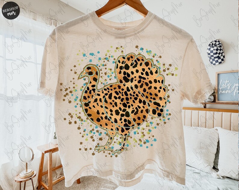 May include: A cream-colored t-shirt featuring a turkey design with a leopard print pattern on the body and tail. The turkey is surrounded by teal and brown speckles. The shirt is hanging on a wooden hanger.
