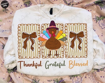 Könnte beinhalten: Weißes Langarm-Sweatshirt mit einem Thanksgiving-Truthahn-Design. Der Truthahn trägt einen Hut und hat bunte Federn. Die Worte "Thankful, Grateful, Blessed" sind unten aufgedruckt. Zwei braune Schleifen flankieren den Truthahn.
