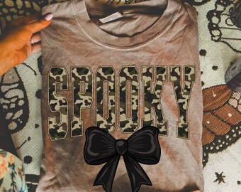 May include: A brown t-shirt with the word "SPOOKY" in camouflage lettering and a black bow graphic. The shirt is folded and displayed on a patterned surface. The shirt is a comfortable, casual style.