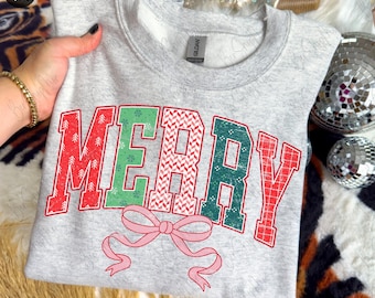Può includere: Felpa grigio chiaro con la scritta "MERRY" a blocchi rossi e verdi. Le lettere presentano motivi festivi e sono decorate con un fiocco rosa. Un capo d'abbigliamento a tema festivo.
