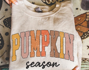 Puede incluir: Una sudadera beige con las palabras "PUMPKIN season" en una fuente retro y desgastada. El texto está en tonos de naranja, rosa y marrón. La sudadera está doblada y sostenida por manos.