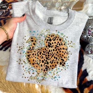 May include: A light gray sweatshirt featuring a turkey design with a leopard print pattern. The turkey is outlined in black and has brown and black spots. The design includes gold, blue, and brown speckles.